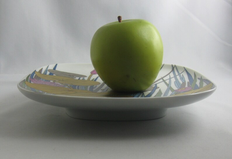 May include: A white ceramic serving dish with a green apple on top. The dish has a floral design with blue, green, yellow, and pink accents.