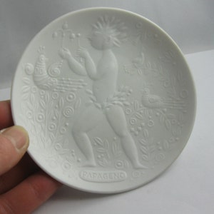 May include: White porcelain plate with a raised design of a figure with a bird and floral motifs. The plate is inscribed with the word "Papageno".