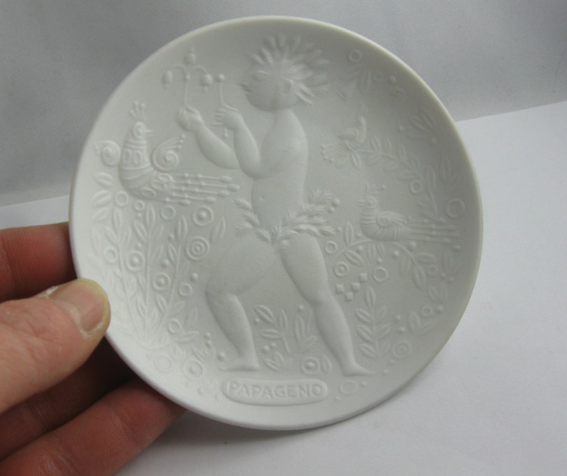 May include: White porcelain plate with a raised design of a figure with a bird and floral motifs. The plate is inscribed with the word "Papageno".