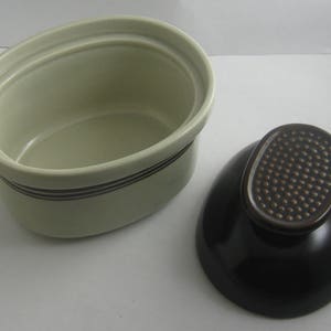 May include: A white oval ceramic dish with a brown rim and a black lid. The lid has a textured brown knob.