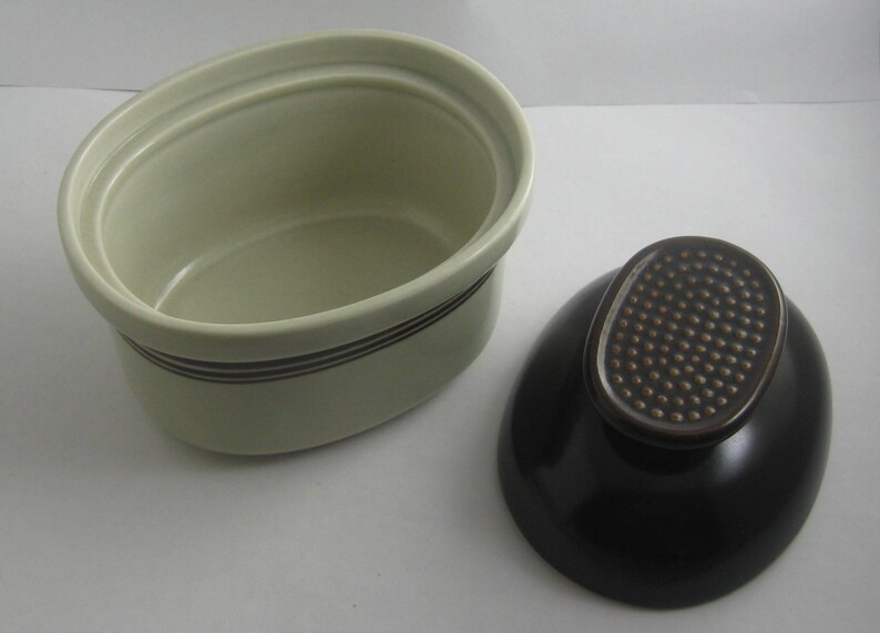 May include: A white oval ceramic dish with a brown rim and a black lid. The lid has a textured brown knob.