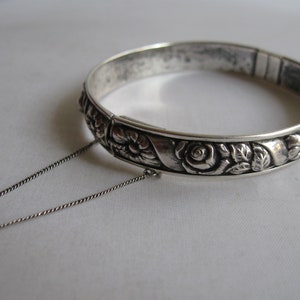May include: A silver bangle bracelet with a floral design. The bracelet has a delicate chain attached to it.