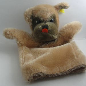 May include: A brown plush hand puppet shaped like a dog with a red nose and black eyes. The puppet is sitting inside a brown plush pouch.