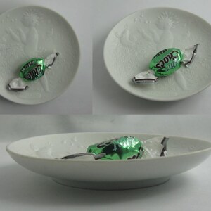 May include: Three white ceramic plates with a raised design of a woman holding a bird. Each plate has a green and silver wrapped mint chocolate candy on top.