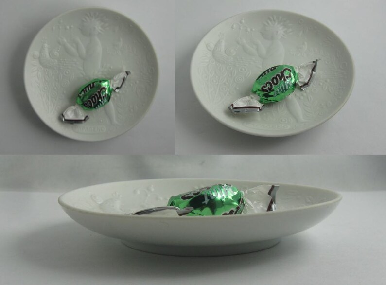 May include: Three white ceramic plates with a raised design of a woman holding a bird. Each plate has a green and silver wrapped mint chocolate candy on top.