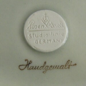 May include: A white porcelain plate with a round, white label that reads "Rosenthal Studio-Linie GERMANY". Below the label is the text "Handgemalt" in brown.