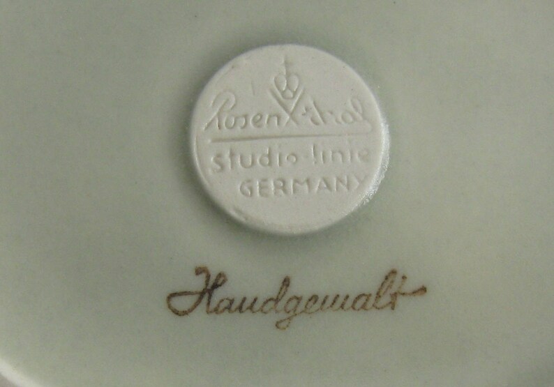 May include: A white porcelain plate with a round, white label that reads "Rosenthal Studio-Linie GERMANY". Below the label is the text "Handgemalt" in brown.