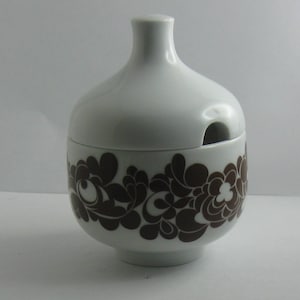 May include: White ceramic sugar bowl with a lid and a brown floral pattern. The lid has a small opening for pouring.