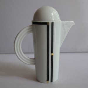 May include: White ceramic creamer with a black and gold geometric design. The creamer has a rounded lid and a curved handle.