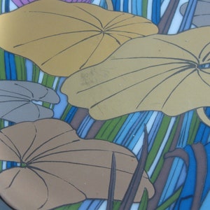 May include: A ceramic plate with a blue, green, and brown striped background. The plate features a design of overlapping, stylized leaves in shades of brown, gray, and gold.