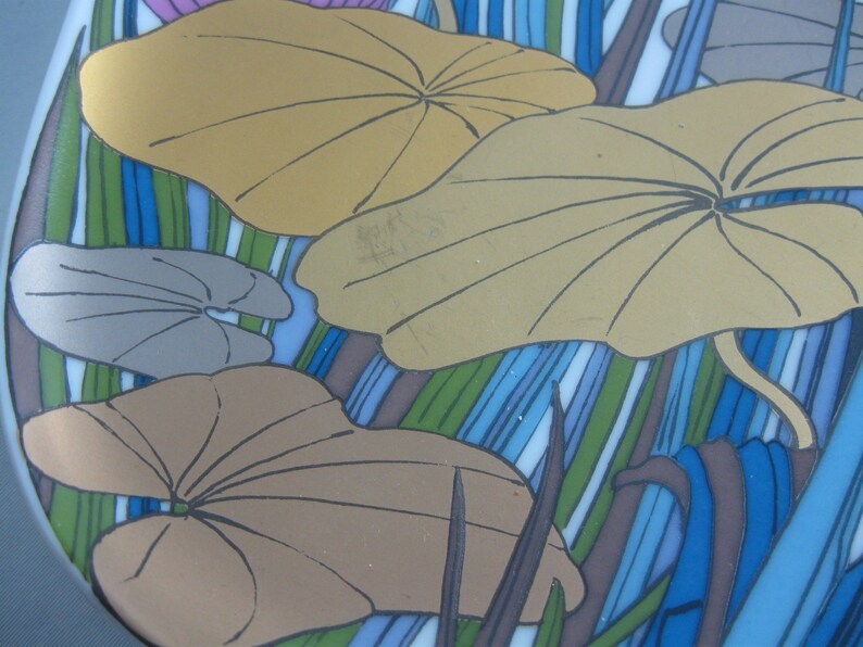 May include: A ceramic plate with a blue, green, and brown striped background. The plate features a design of overlapping, stylized leaves in shades of brown, gray, and gold.