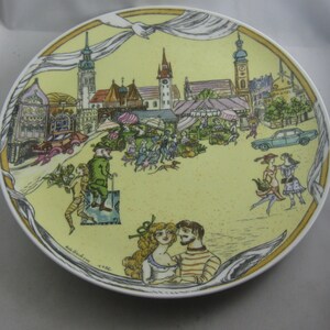 May include: A decorative plate with a yellow background featuring a detailed illustration of a European town square with people, cars, and market stalls. The plate has a white rim and a gold border.