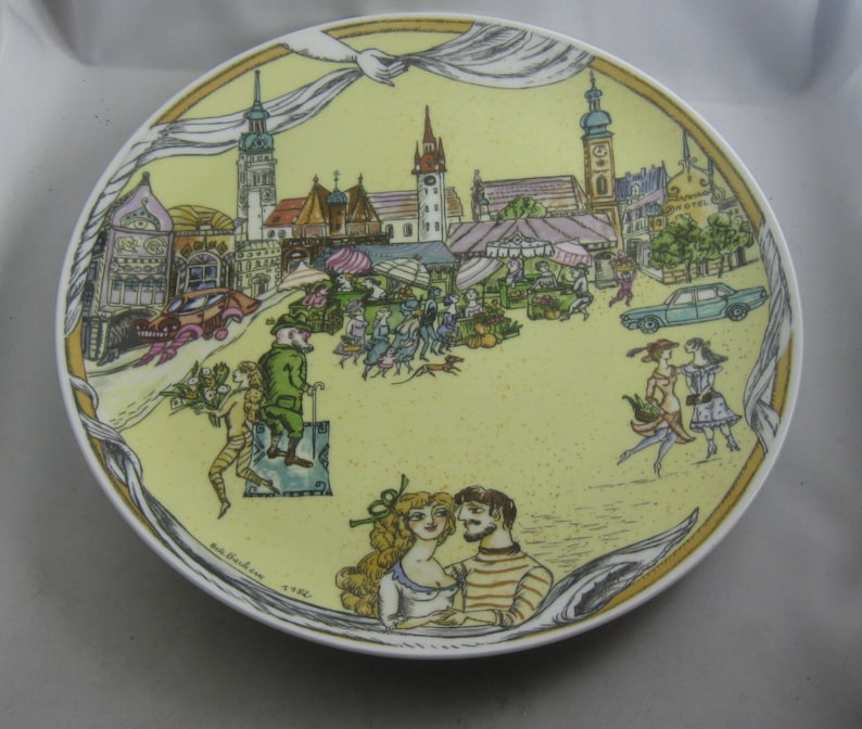 May include: A decorative plate with a yellow background featuring a detailed illustration of a European town square with people, cars, and market stalls. The plate has a white rim and a gold border.