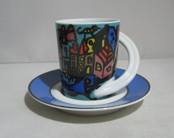 May include: A white ceramic coffee cup with a blue and white saucer. The cup features a whimsical design of a city with colourful buildings and a unique curved handle.