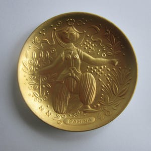 Puede incluir: Un plato de metal dorado con un diseño en relieve de una mujer con una túnica fluida, rodeada de hojas y flores estilizadas. El plato está inscrito con la palabra "PAMINA".