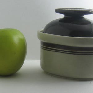 May include: A white ceramic jar with a brown stripe design and a black lid. The jar is sitting on a white surface next to a green apple.