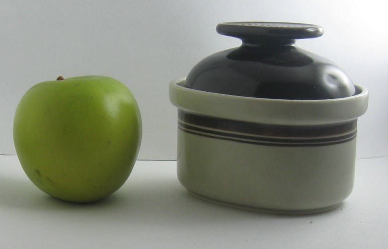May include: A white ceramic jar with a brown stripe design and a black lid. The jar is sitting on a white surface next to a green apple.