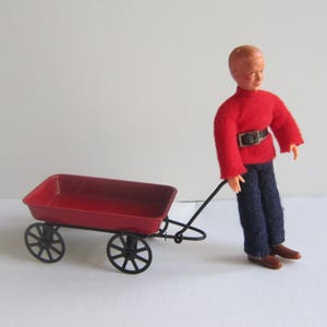 May include: A red toy wagon with black wheels and a black handle. A doll with a red shirt and blue pants is pulling the wagon.