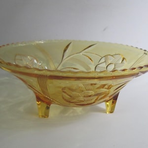 May include: A yellow glass bowl with a floral design and three legs. The bowl has a scalloped edge and is made of clear glass.