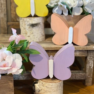 Pu&ograve; includere: Tre figurine di farfalla in legno, dipinte in giallo, pesca e viola, su basi in legno di betulla.