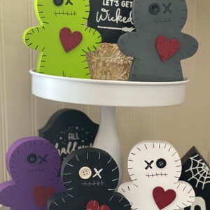 May include: A collection of wooden voodoo doll figurines in various colors, including green, gray, purple, black, and white. Each doll has stitched details, a heart, and a button eye. The phrase "LET'S GET Wicked" is on a black sign.