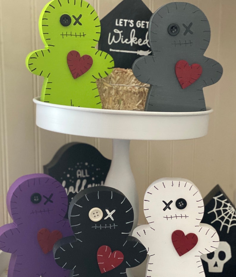May include: A collection of wooden voodoo doll figurines in various colors, including green, gray, purple, black, and white. Each doll has stitched details, a heart, and a button eye. The phrase "LET'S GET Wicked" is on a black sign.