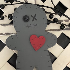 May include: A gray wooden doll with stitched details, a button eye, and an 'X' for a mouth. A red heart is stitched onto the doll's chest. The doll is set against a black and white checkered background, with black berry garland.