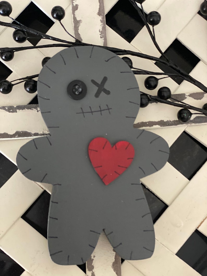 May include: A gray wooden doll with stitched details, a button eye, and an 'X' for a mouth. A red heart is stitched onto the doll's chest. The doll is set against a black and white checkered background, with black berry garland.