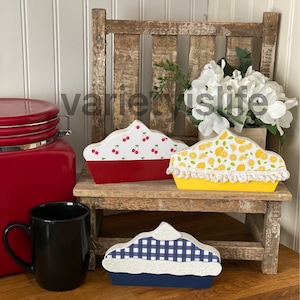 May include: Three wooden pie-shaped decorations with different designs. The first pie is red with white cherries, the second is yellow with lemons, and the third is blue with a white and blue checkered pattern. The pies are sitting on a wooden bench.