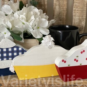 May include: Three wooden pie slice decorations. The first is blue and white gingham with a white top. The second is yellow and white with a white top. The third is red and white with a cherry pattern and a white top.