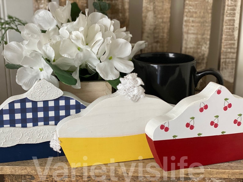 May include: Three wooden pie slice decorations. The first is blue and white gingham with a white top. The second is yellow and white with a white top. The third is red and white with a cherry pattern and a white top.