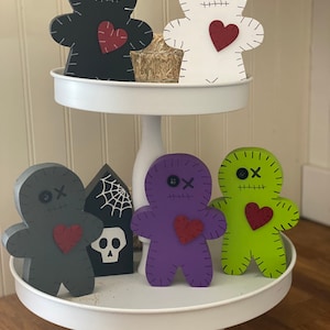 May include: A two-tiered white serving tray displays six wooden voodoo doll figures in black, white, gray, purple, and green. Each doll has a red heart, stitched details, and button eyes. A small tombstone with a spiderweb and skull is also present.