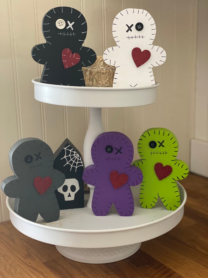 May include: A two-tiered white serving tray displays six wooden voodoo doll figures in black, white, gray, purple, and green. Each doll has a red heart, stitched details, and button eyes. A small tombstone with a spiderweb and skull is also present.