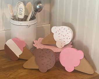 Puede incluir: Cuatro decoraciones de cono de helado de madera con pintura rosa, blanca y marrón. Los conos tienen chispas en la parte superior y están dispuestos sobre una superficie de madera.