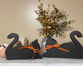 May include: Two black cat silhouettes crafted from wood, each featuring a curved tail and a decorative orange ribbon tied around the neck. The ribbons are patterned with small black dashes, adding a subtle detail to the solid black cat figures.