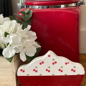 May include: A white wooden pie shaped decorative piece with a red cherry pattern and a red base. The piece is sitting on a wooden surface next to a red ceramic canister.