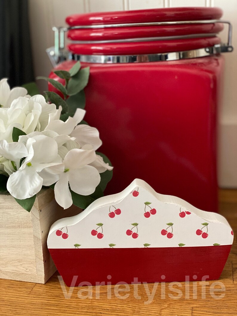 May include: A white wooden pie shaped decorative piece with a red cherry pattern and a red base. The piece is sitting on a wooden surface next to a red ceramic canister.