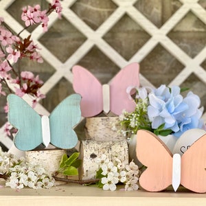Pu&ograve; includere: Tre decorazioni in legno a forma di farfalla nei colori rosa, azzurro e pesca. Le farfalle sono dipinte con dettagli bianchi e sono esposte su una superficie in legno con uno sfondo a traliccio bianco.