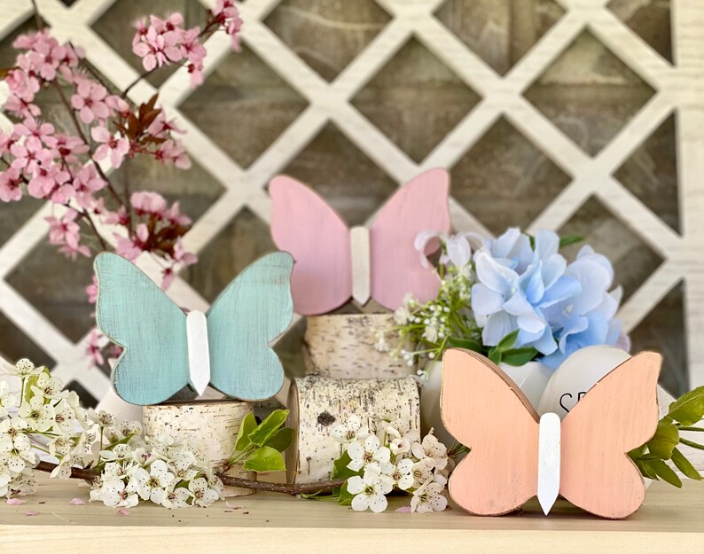 Pu&ograve; includere: Tre decorazioni in legno a forma di farfalla nei colori rosa, azzurro e pesca. Le farfalle sono dipinte con dettagli bianchi e sono esposte su una superficie in legno con uno sfondo a traliccio bianco.