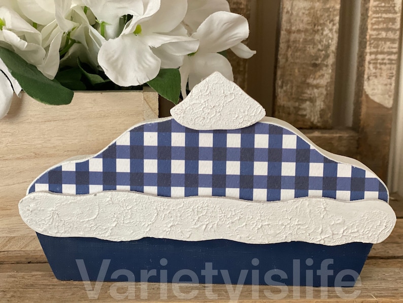 May include: A blue and white checkered pie crust with a white, textured top. The pie crust is made of wood and is a decorative piece.