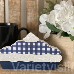 May include: A blue and white checkered pie with a white crust and a blue base. The pie is made of wood and is a decorative piece.