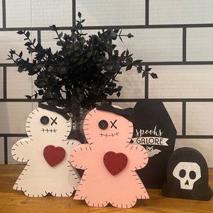 May include: Wooden Halloween decorations including a white and pink voodoo doll with a red heart, a black coffin with "spooks galore" text, and a black skull. The dolls have button eyes and stitched details.
