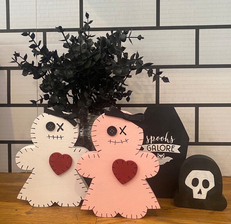 May include: Wooden Halloween decorations including a white and pink voodoo doll with a red heart, a black coffin with "spooks galore" text, and a black skull. The dolls have button eyes and stitched details.