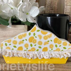 May include: A yellow wooden lemon pie shaped mug holder with white crochet trim and a lemon print. The holder is sitting on a wooden surface with a black mug and white flowers in a wooden planter.