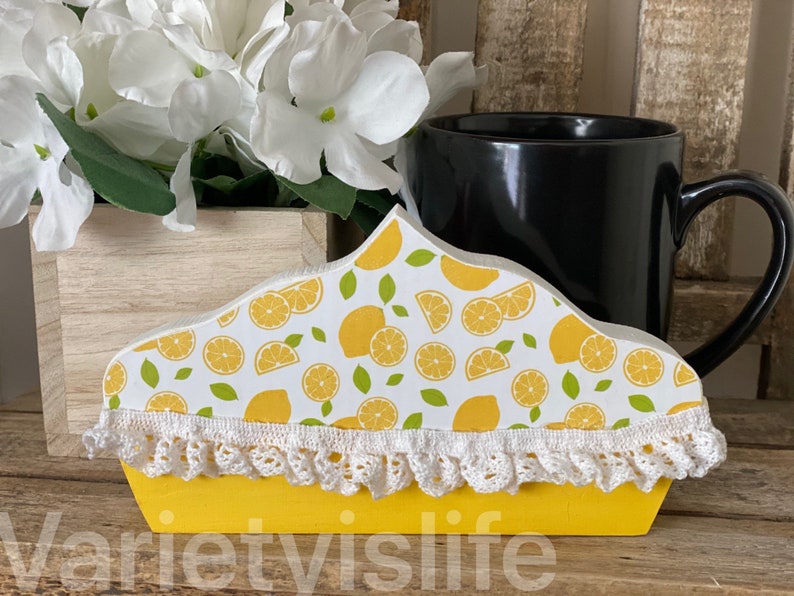 May include: A yellow wooden lemon pie shaped mug holder with white crochet trim and a lemon print. The holder is sitting on a wooden surface with a black mug and white flowers in a wooden planter.