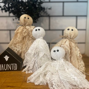 May include: Four ghost-shaped ornaments made of white and beige lace fabric with round heads and black dot eyes. A small black wooden house-shaped sign with a white ghost icon and the text HAUNTED is positioned next to the lace ghost figures.