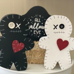 May include: Two wooden Halloween-themed figures. One is black, the other white, both shaped like gingerbread men with stitched details, red hearts, and button eyes. A black sign behind them reads "ALL HALLOWS EVE".