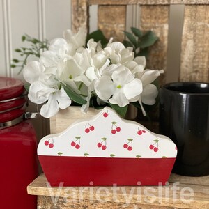 May include: A wooden cherry pie-shaped napkin holder with a red base and a white top with a cherry pattern. The holder is sitting on a wooden shelf with a white flower arrangement in the background.