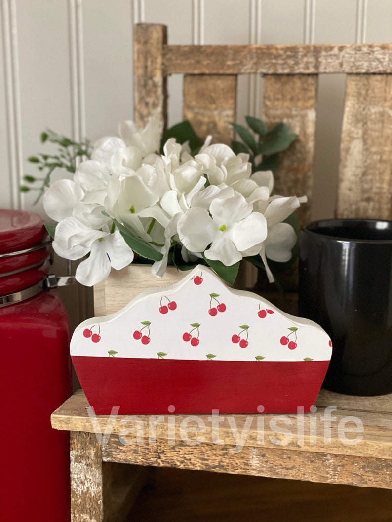 May include: A wooden cherry pie-shaped napkin holder with a red base and a white top with a cherry pattern. The holder is sitting on a wooden shelf with a white flower arrangement in the background.