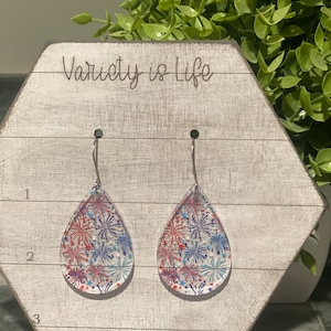 May include: A pair of teardrop-shaped earrings with a clear background and a red, white, and blue firework pattern. The earrings are hanging on a white wooden display with the text "Variety is Life" written on it.